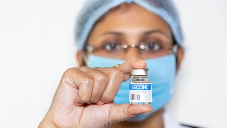 Indischer Arzt in chirurgischer Maske hält eine Impfstoffflasche in die Kamera.