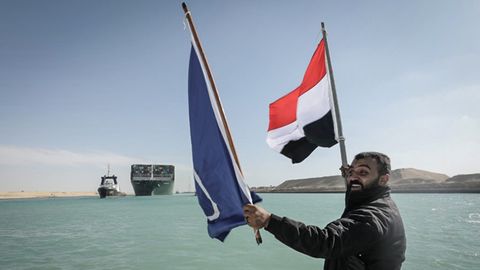 Ein Arbeiter schwenkt eine Flagge Ägyptens, im Hintergrund das Containerschiff "Ever Given" 