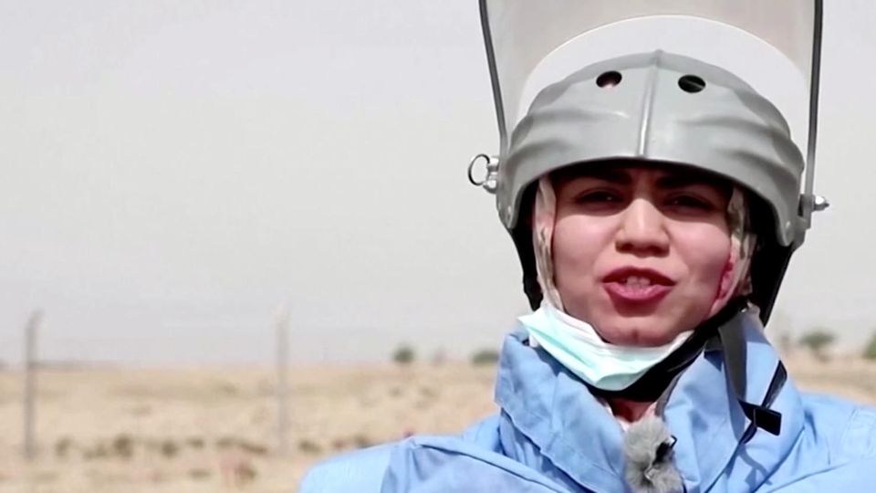 Eine Frau steht in hellblauer Sprengmeister-Schutzausrüstung im Freien in der Sonne