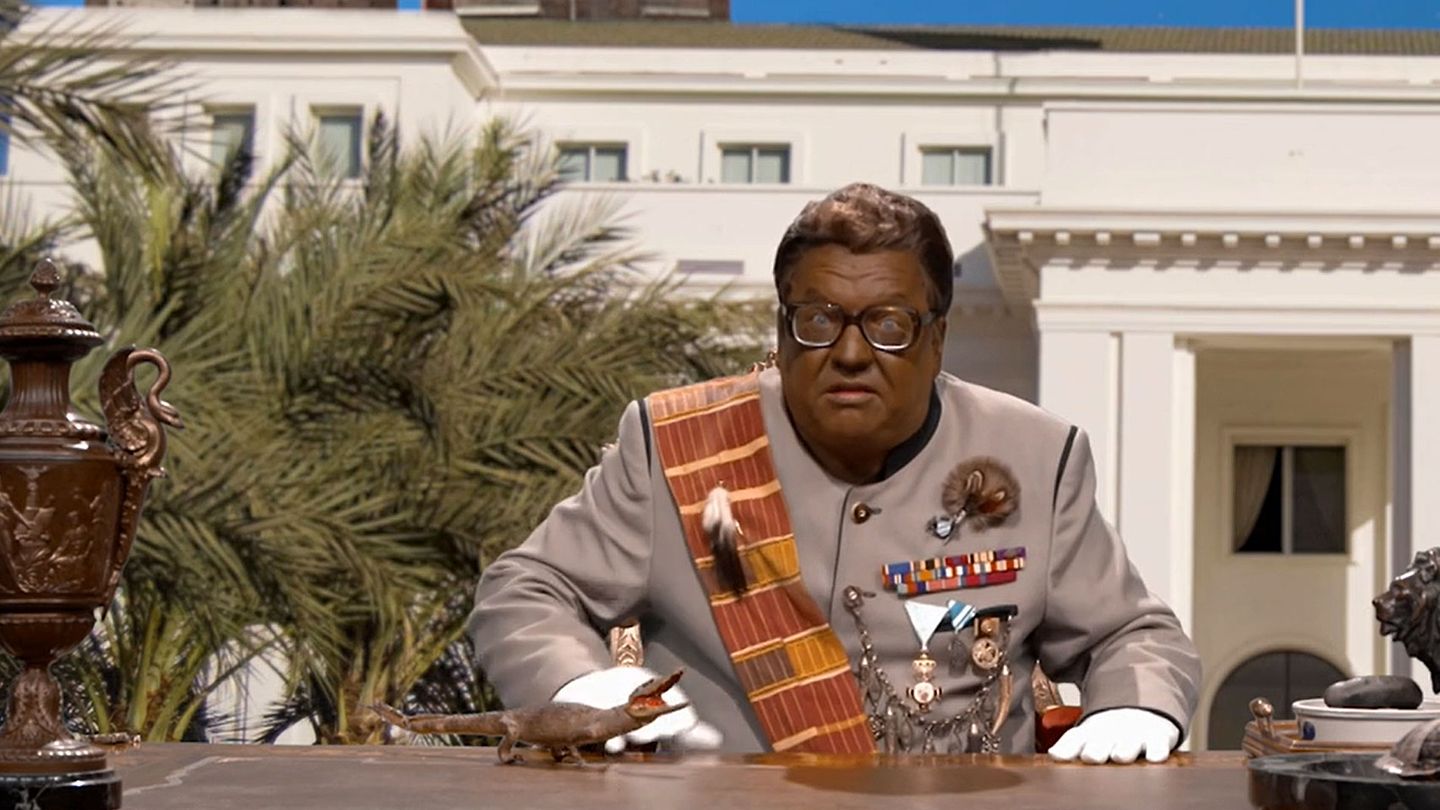 Satiriker Helmut Schleich sitzt als afrikanischer Despot mit schwarz angemaltem Gesicht am Schreibtisch