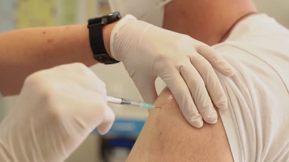 Hände in Gummihandschuhen stechen eine Spritze in den linken Oberarm eines Mannes