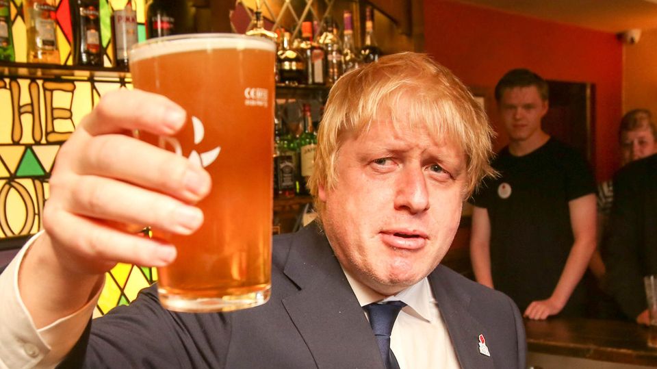 Ein blonder, propperer Mann im Anzug erhebt ein Pint-Glas mit Bier, während er in einem englischen Pub steht