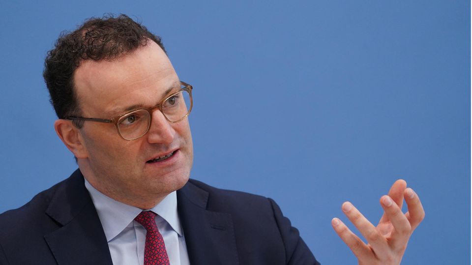 Ein Mann mit braunen Locken und durchsichtigem Brillengestell sitzt im Anzug vor einer blauen Wand und gestikuliert mit links