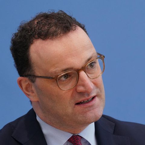 Ein Mann mit braunen Locken und durchsichtigem Brillengestell sitzt im Anzug vor einer blauen Wand und gestikuliert mit links
