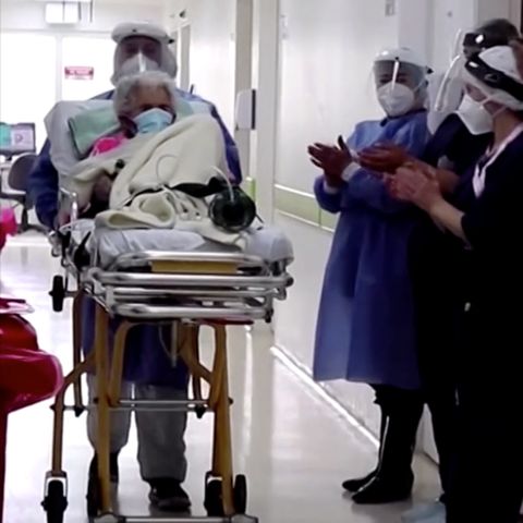 Pflegekräfte in Schutzkleidung schieben eine weißhaarige Frau auf einer Liege aus einem Krankenhaus heraus