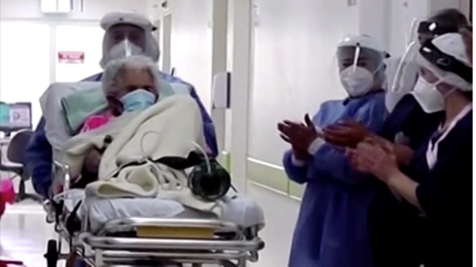 Pflegekräfte in Schutzkleidung schieben eine weißhaarige Frau auf einer Liege aus einem Krankenhaus heraus