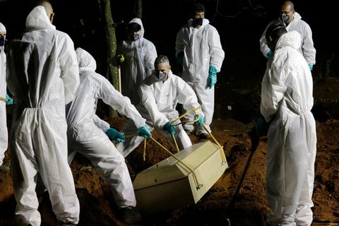 Sieben Männer in weißen Schutzanzügen senken einen hellen Holzsarg mit einem Corona-Toten in ein Grab