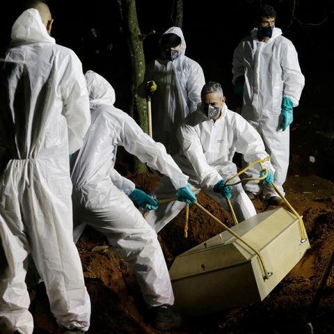 Sieben Männer in weißen Schutzanzügen senken einen hellen Holzsarg mit einem Corona-Toten in ein Grab