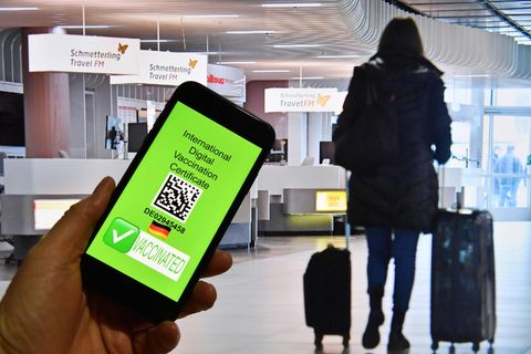 Eine Hand hält ein Handy, dahinter ist eine Szene im Flughafen zu sehen