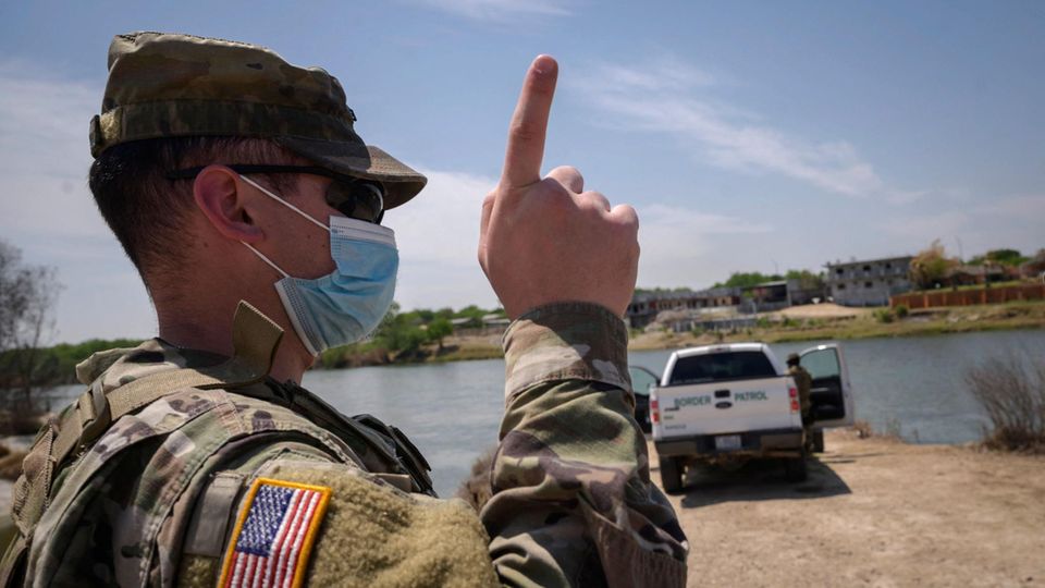 USA Border Patrol Grenze Mexiko Migration Ein Uniformierter mit US-Flagge auf dem rechten Ärmel steht in einer Wüste und hebt den Zeigefinger. Im Hintergrund ein Pickup