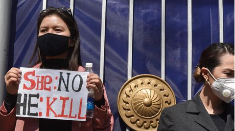 Eine Frau steht mit Maske und Schild vor einem Tor. Auf dem Schild steht: "She: no - He: kill"