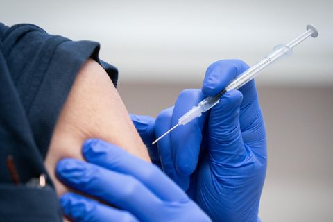 Hände in blauen Gummihandschuhen halten eine aufgezogene Spritze kurz vor einen linken, weißen Oberarm