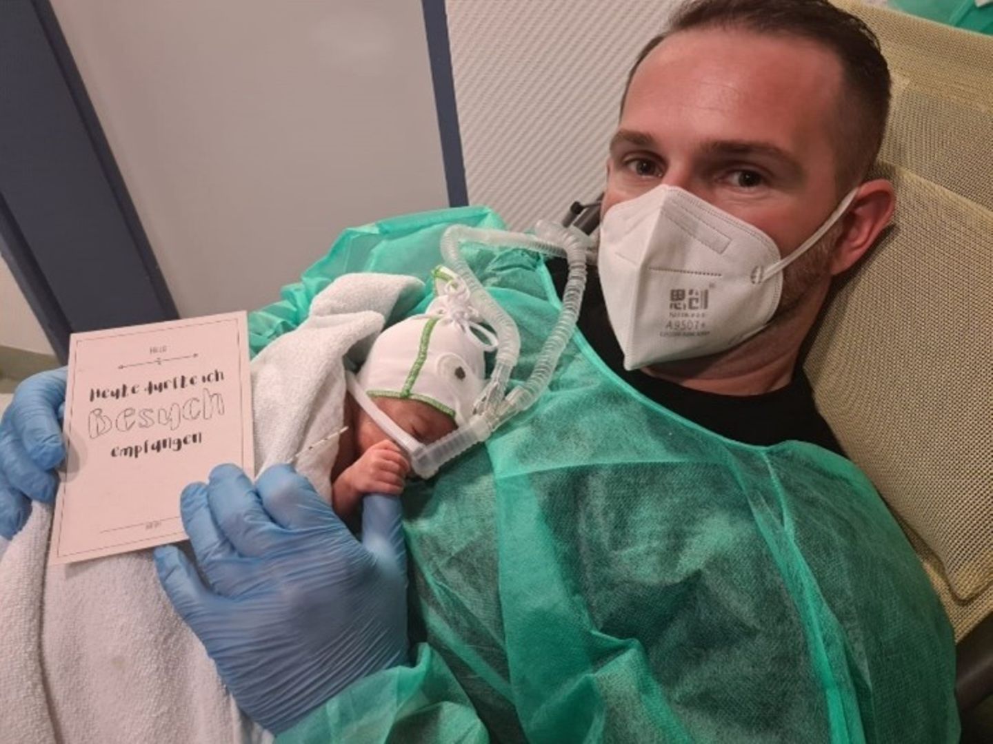 Ein Vater mit FFP2-Maske und Kittel hat seinen kleinen Sohn auf dem Bauch