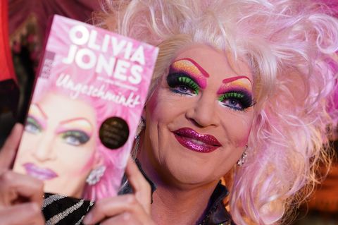 Dragqueen Olivia Jones trägt eine blonde Perücke mit rosa Strähnen und hält ihr Buch "Ungeschminkt" neben ihr Gesicht