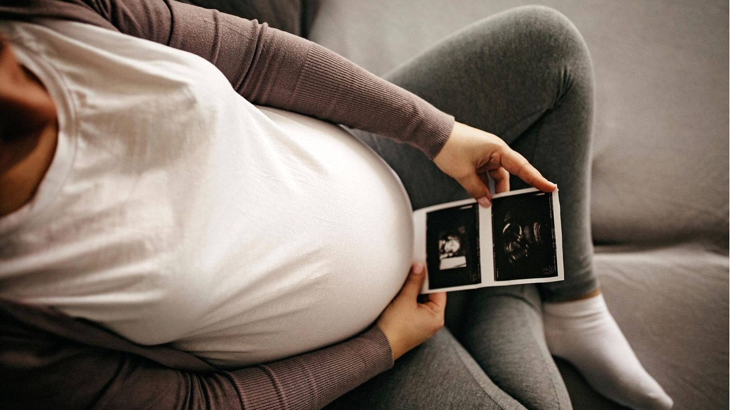 Schwangere sitzt auf dem Sofa und schaut auf das Ultraschallbild ihres Babys, das sie vor ihren runden Bauch hält