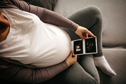 Schwangere sitzt auf dem Sofa und schaut auf das Ultraschallbild ihres Babys, das sie vor ihren runden Bauch hält