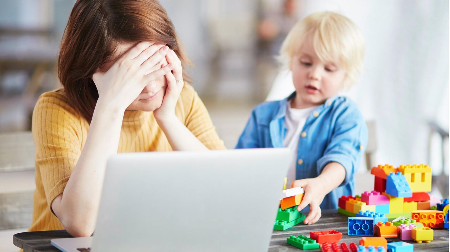 Erschöpfung durch die Pandemie Frau sitzt vor genervt und erschöpft vor dem Laptop, während ihr Kind neben ihr auf dem Tisch mit LEGO-Steinen spielt