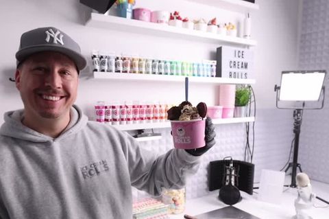 Ein junger Mann in grauem Kapuzenpulli und "NY"-Cap lächelt, während er mit links einen vollen rosa Papp-Eisbecher hochhält