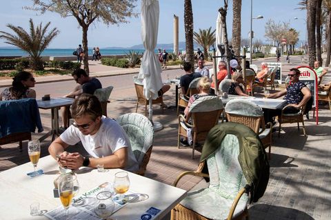 Auf der Terrasse eines Restaurant sind alle Tische besetzt. Die Gäste sitzen in der Sonne. Hinter ihnen Palmen, Strand und Meer