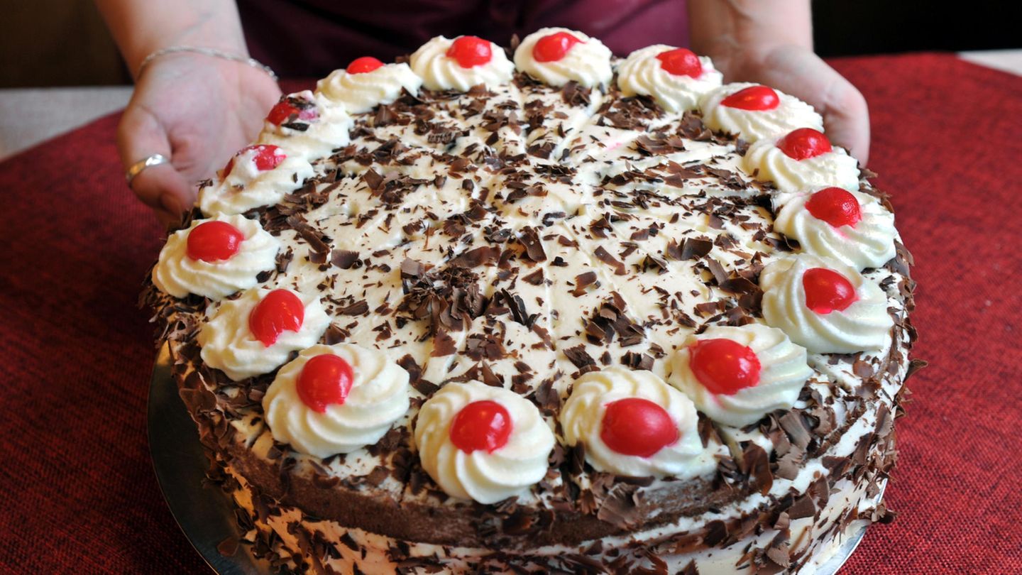 Die Hände einer Frau stellen einen runden, weißen Kuchenteller mit einer Schwarzwälder Kirschtorte auf eine rote Tischdecke