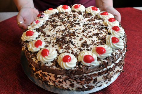 Die Hände einer Frau stellen einen runden, weißen Kuchenteller mit einer Schwarzwälder Kirschtorte auf eine rote Tischdecke