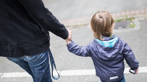 Ein Mann in Jeans und schwarzer Jacke ist mit seinem blonden Kind an der Hand von hinten zu sehen