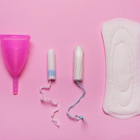 Eine Menstruationstasse, zwei Tampons und eine Binde vor einem pinken Hintergrund