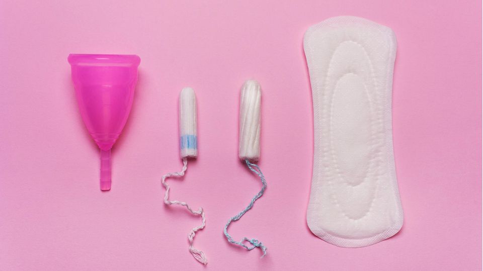Eine Menstruationstasse, zwei Tampons und eine Binde vor einem pinken Hintergrund