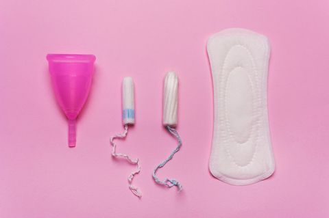 Eine Menstruationstasse, zwei Tampons und eine Binde vor einem pinken Hintergrund