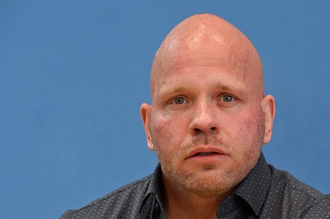 Ein muskulöser, glatzköpfiger weißer Mann sitzt in grauem Hemd mit hellen Punkten vor einer blauen Wand