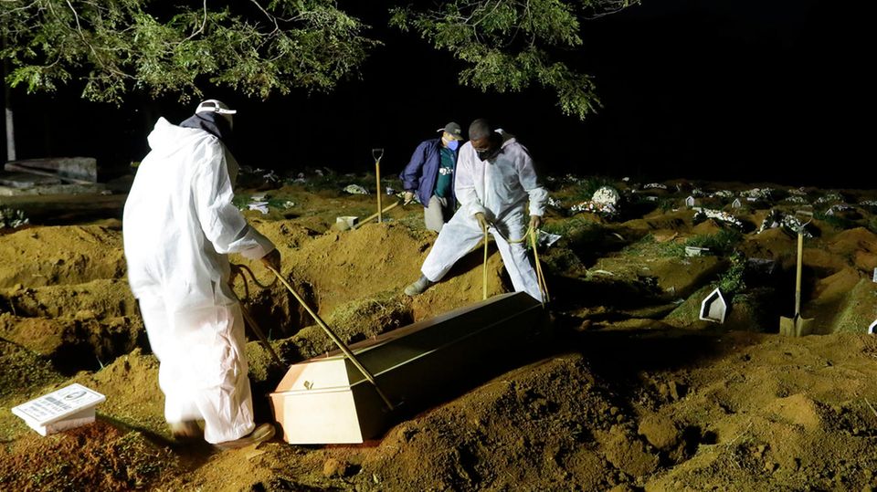 Zwei Menschen in weißen Schutzanzügen senken auf einem dunklen Friedhof einen hellen Holzsarg in eine Grube