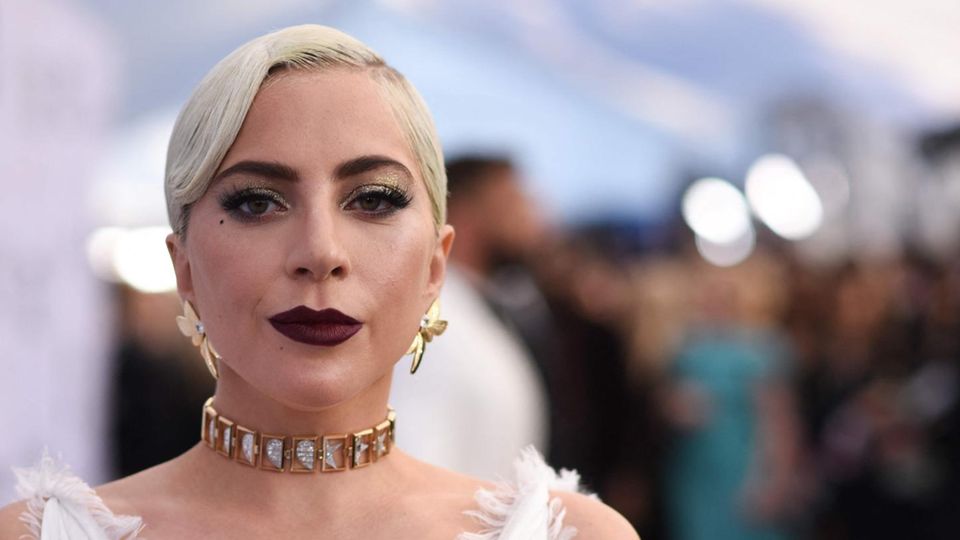 Lady Gaga Eine weiße, blonde Frau in weißem Abendkleid und mit goldenen Ohrringen schaut ernst in die Kamera