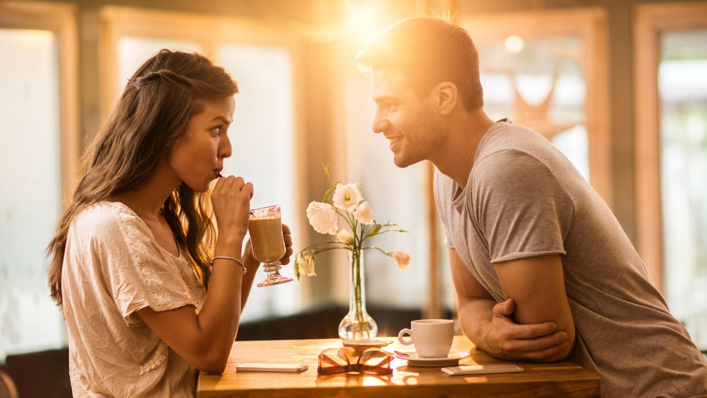 Eine junge Frau und ein junger Mann beim Date im Café. Sie trinkt aus dem Strohhalm, während er sich über den Tisch vorbeugt.