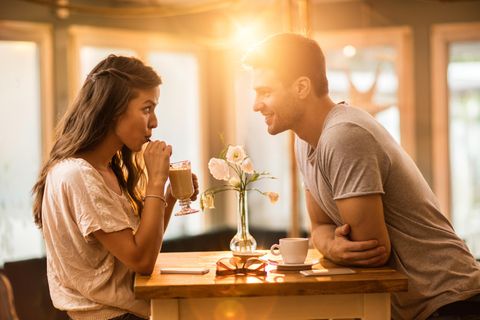 Eine junge Frau und ein junger Mann beim Date im Café. Sie trinkt aus dem Strohhalm, während er sich über den Tisch vorbeugt.