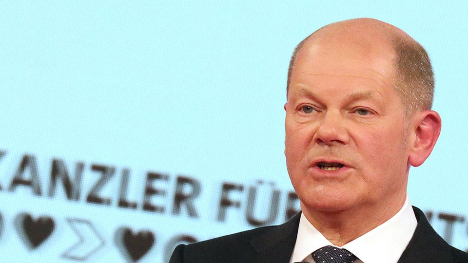 Ein weißer Mann mit kurzrasiertem Haarkranz spricht in Anzug und Krawatte in ein kleines schwarzes Mikrofon