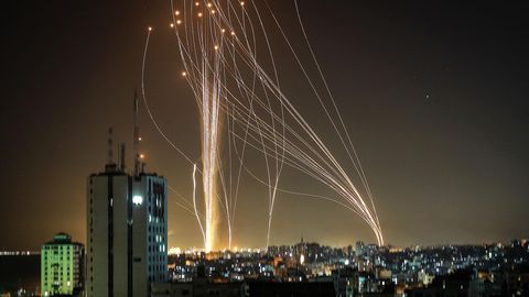 Durch den Nachthimmel über einer Stadt ziehen sich Lichtspuren von Raketen, die von zwei Orten am Horizont starten