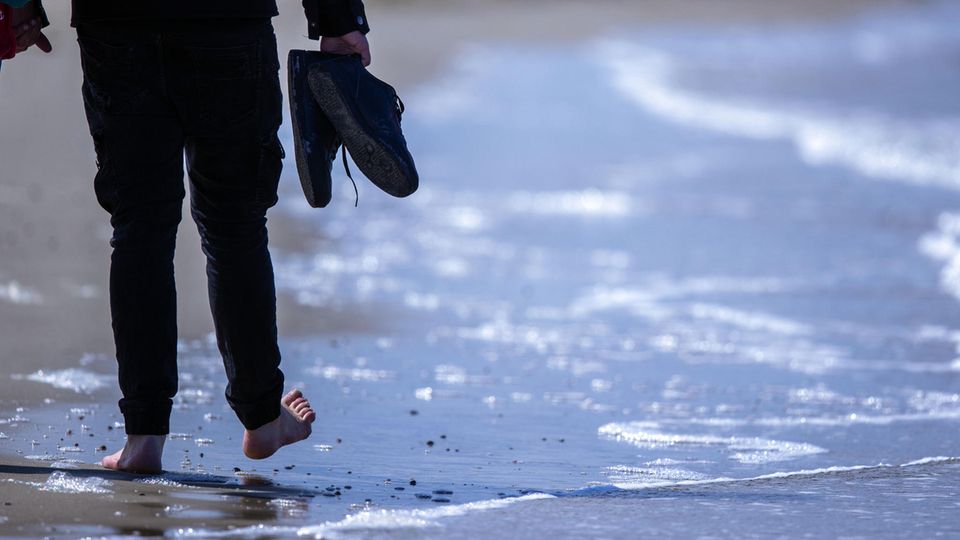 Corona Mecklenburg-Vorpommern Tourismus Von hinten sind die Beine eines Mannes zu sehen, der barfuß am Meeressaum entlang geht und die Schuhe dabei in der Hand hält