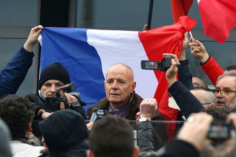 Ein älterer weißer Mann mit sehr kurzen Haaren spricht zu Demonstranten – hinter ihm eine französische Flagge