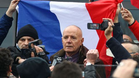 Ein älterer weißer Mann mit sehr kurzen Haaren spricht zu Demonstranten – hinter ihm eine französische Flagge