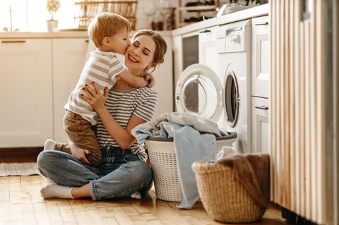 Mutter und Kind sitzen vor der Waschmaschine. Der Sohn drückt seiner Mutter einen Kuss auf die Wange.