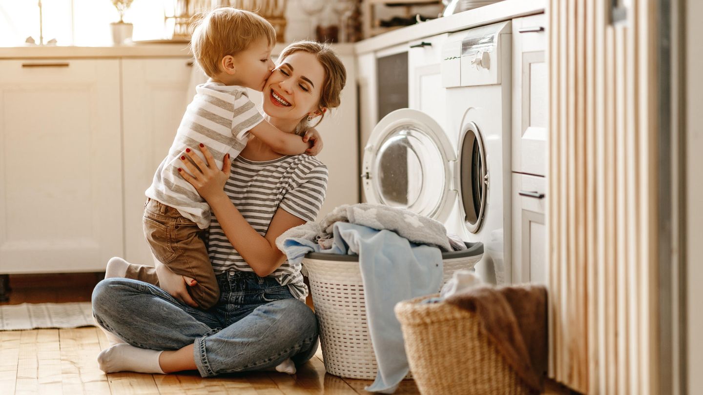 Mutter und Kind sitzen vor der Waschmaschine. Der Sohn drückt seiner Mutter einen Kuss auf die Wange.