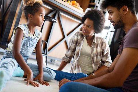 Familie sitzt af dem Boden. Die Eltern erklären der Tochter etwas, die aufmerksam zuhört.
