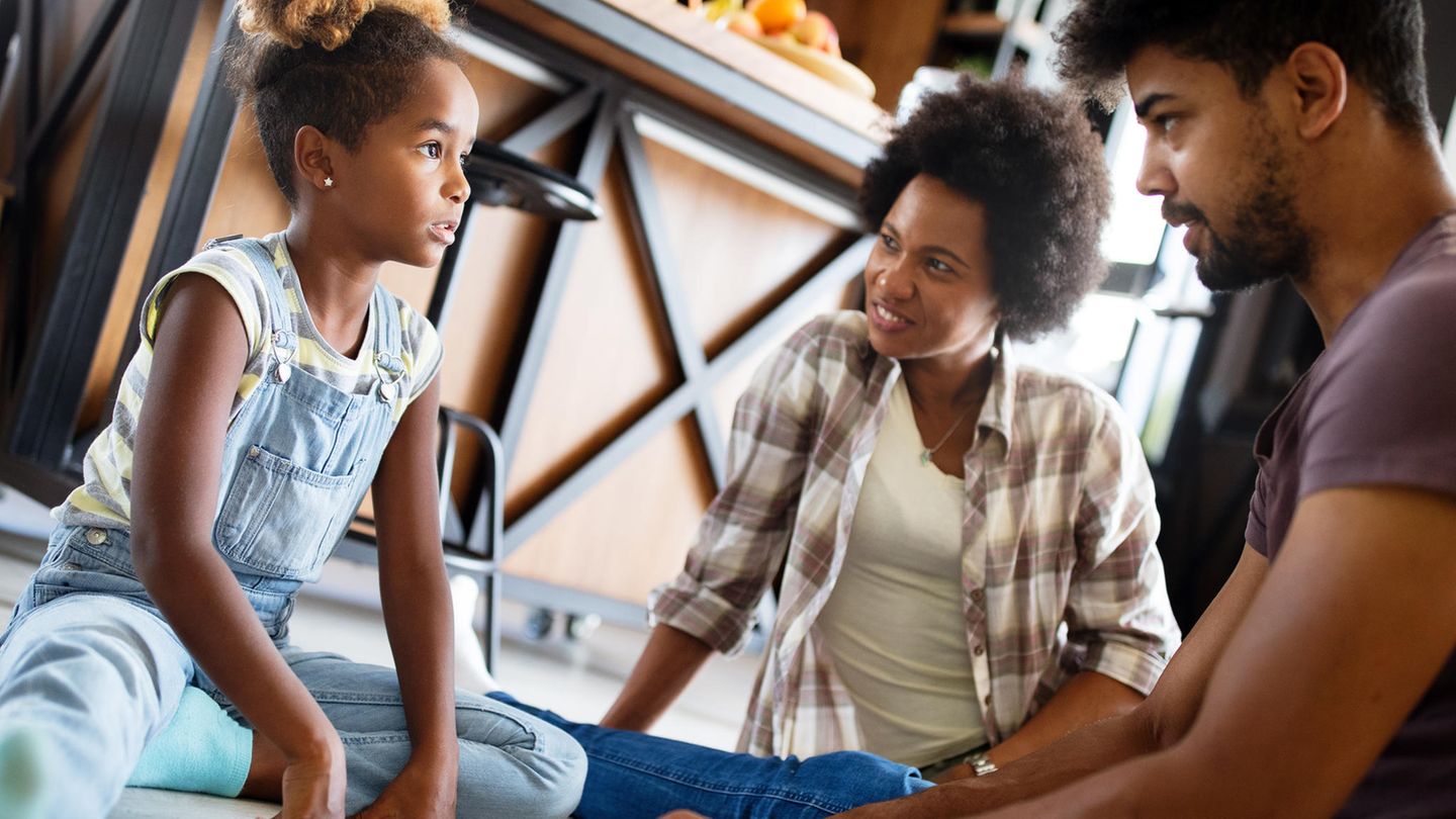 Familie sitzt af dem Boden. Die Eltern erklären der Tochter etwas, die aufmerksam zuhört.