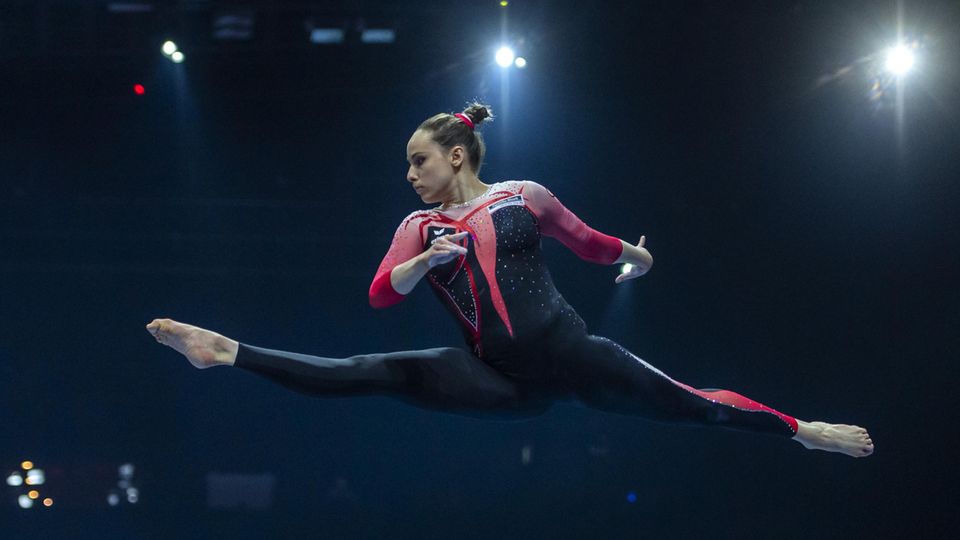 Sarah Voss Turnerin Sarah Voss springt im Spagatsprung auf dem Balken und trägt dabei einen langen Anzug