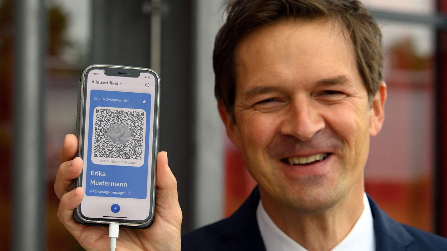 Ein weißer Mann im blauen Jackett lächelt, während er ein Smartphone-Display mit blau umrandetem QR-Code hochhält