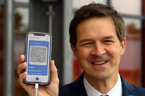 Ein weißer Mann im blauen Jackett lächelt, während er ein Smartphone-Display mit blau umrandetem QR-Code hochhält