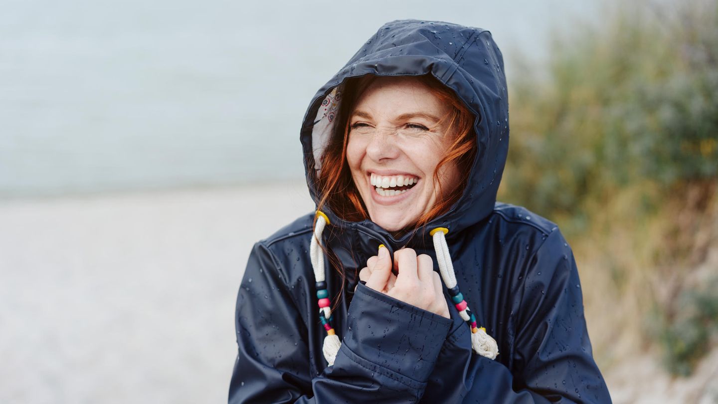 Lachende Frau mit roten Haaren im Regenmantel vor dem Meer