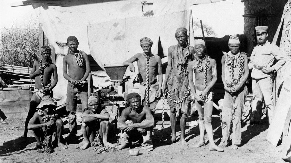 Ein Schwarz-Weiß-Foto zeigt einen mutmaßlich deutschen Uniformierten, der in Ketten gelegte Schwarze bewacht