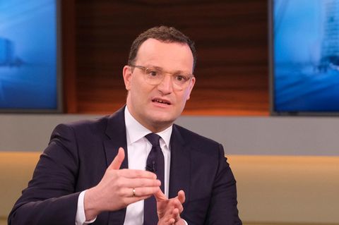 Ein Mann mit Brille und braunen Locken sitzt im Anzug in einem TV-Studio und gestikuliert mit beiden Händen vor der Brust