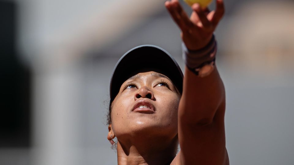 Eine asiatisch aussehende Frau schaut bei Sonnenschein hoch zum Tennisball, den ihre linke Hand gerade hochwirft